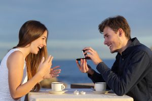 Boyfriend requesting hand of his girlfriend with a engagement ring in a restaurant with the ocean in the background